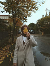 woman dressed modestly in brown hijab cream trench coat on sidewalk leaves falling holding leaf