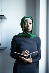 women in green hijab black maxi dress holding binder and pen looking at camera in an office with bookshelves